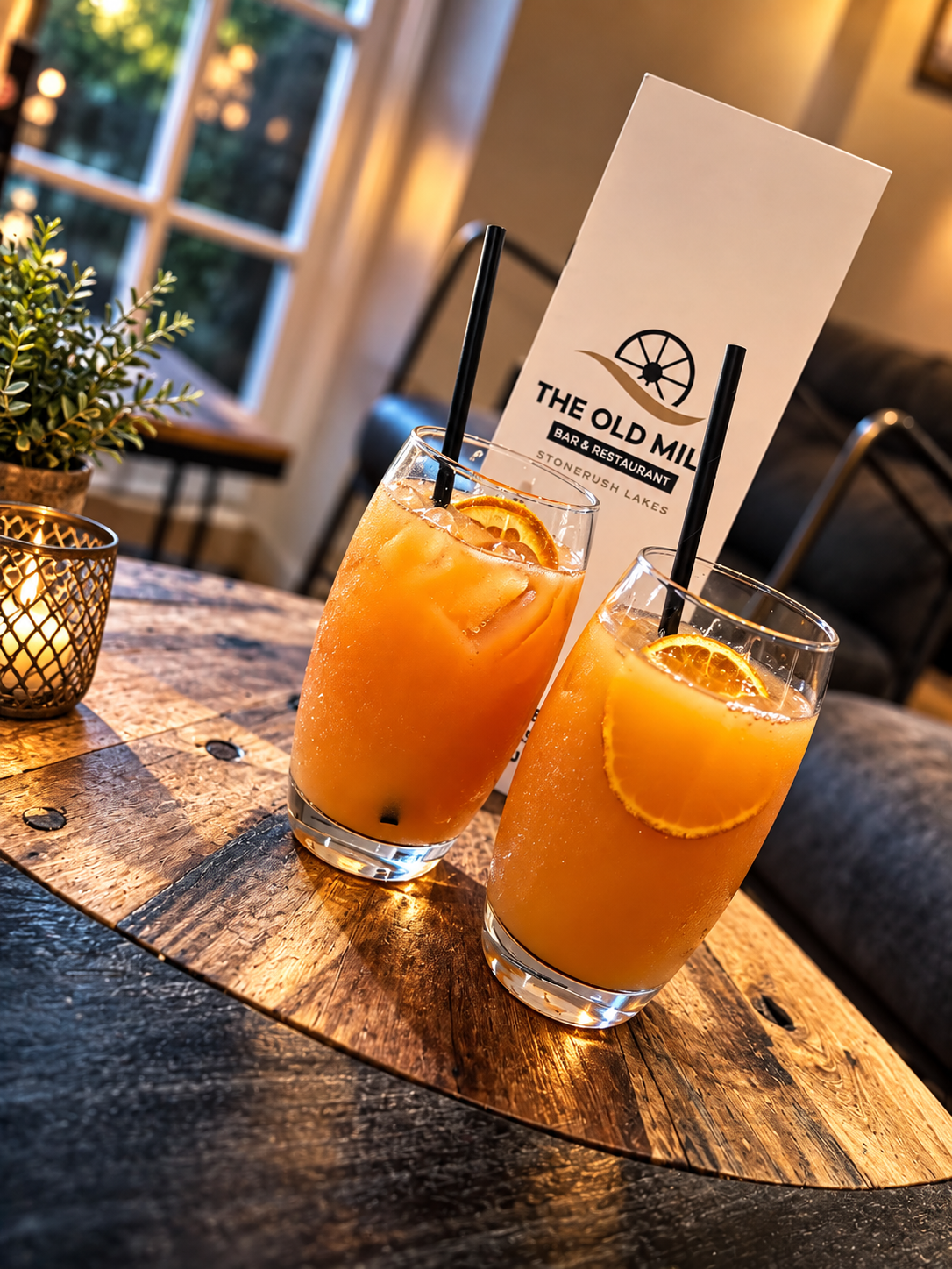 Two fresh orange cocktails in highball glasses with orange slices, beside a candle and menu card